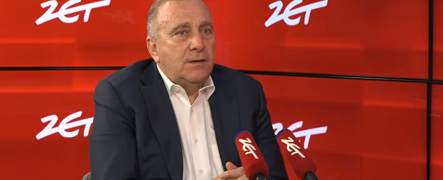 Mężczyzna w garniturze siedzi przy stole, mówi do mikrofonów w studio radiowym. Tło w kolorze czerwonym.