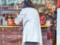 Osoba w białym fartuchu przygotowuje leki w aptece, za nią półki z produktami farmaceutycznymi.