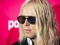 Mężczyzna z długimi, blond włosami i okularami przeciwsłonecznymi na różowym tle.