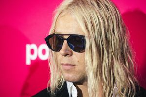 Mężczyzna z długimi, blond włosami i okularami przeciwsłonecznymi na różowym tle.