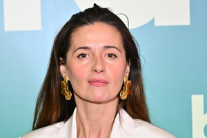 Agnieszka Grochowska z długimi włosami w jasnej bluzce i dużych złotych kolczykach, na tle zielonego tła.