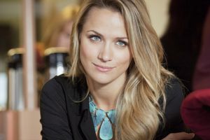 Kobieta z długimi blond włosami, uśmiechnięta, w czarnej bluzce i niebieskiej biżuterii.