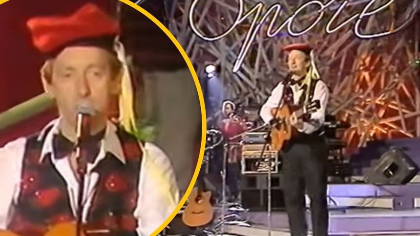 Mężczyzna w czerwonej czapce gra na gitarze, na scenie w tle muzycy i oświetlenie.