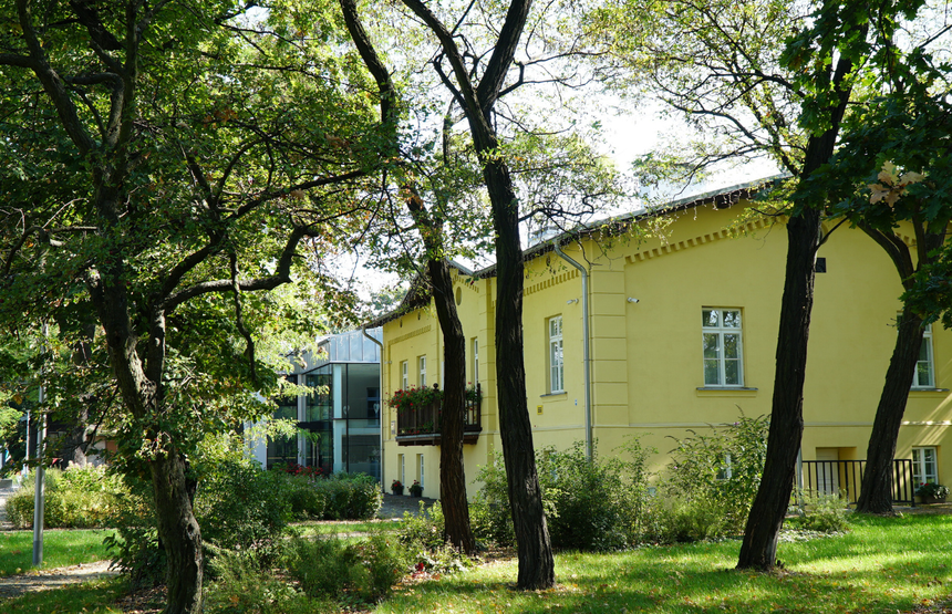 Zielony park z drzewami i żółtym budynkiem w tle, w słoneczny dzień.