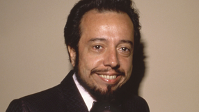 Uśmiechnięty Sergio Mendes w eleganckiej marynarce na jasnym tle.