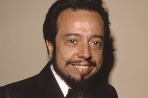 Uśmiechnięty Sergio Mendes w eleganckiej marynarce na jasnym tle.