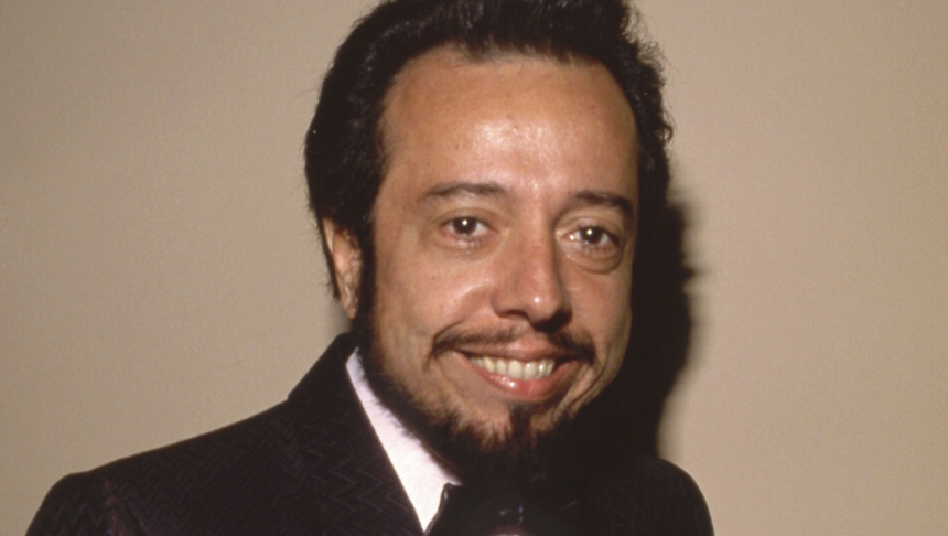 Uśmiechnięty Sergio Mendes w eleganckiej marynarce na jasnym tle.