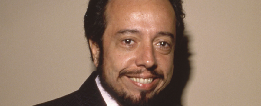 Uśmiechnięty Sergio Mendes w eleganckiej marynarce na jasnym tle.