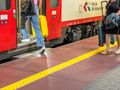 Na stacji, pasażerowie wychodzą z pociągu, wśród nich bagaże na kółkach. Kolorowy pociąg w tle.