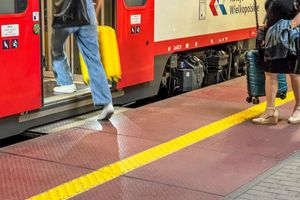 Na stacji, pasażerowie wychodzą z pociągu, wśród nich bagaże na kółkach. Kolorowy pociąg w tle.