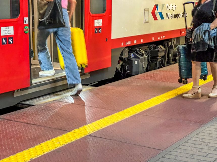 Na stacji, pasażerowie wychodzą z pociągu, wśród nich bagaże na kółkach. Kolorowy pociąg w tle.