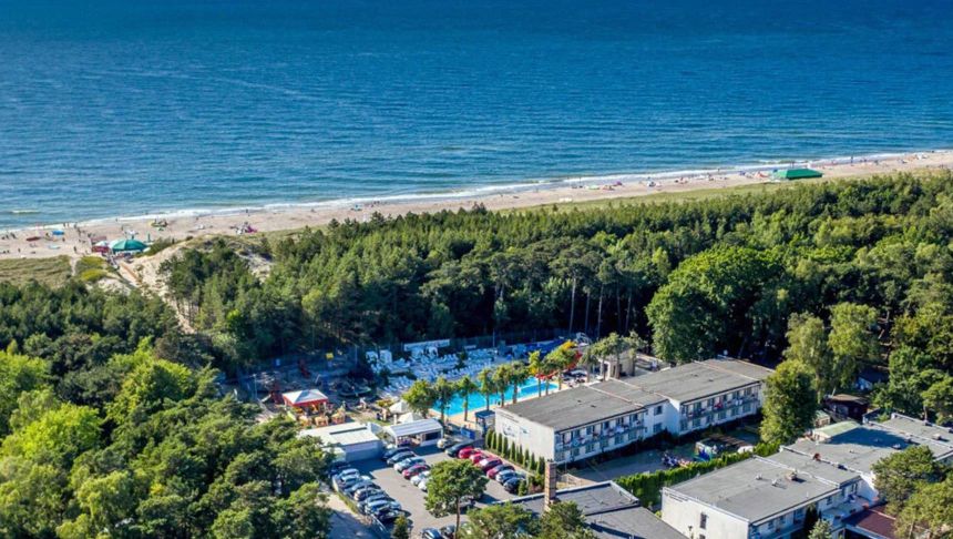 Widok na plażę nadmorską z hotelami, drzewami i basenem. W tle morze i piasek.