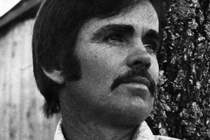 Mężczyzna z wąsami, patrzący w dal, stoi obok drzewa. Czarnobiałe zdjęcie.