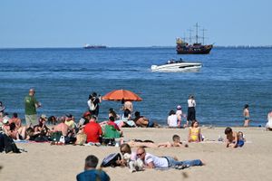 Plaża z wypoczywającymi ludźmi, pływającymi łodziami i statkiem. Jasne niebo, piasek.
