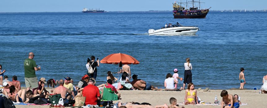 Plaża z wypoczywającymi ludźmi, pływającymi łodziami i statkiem. Jasne niebo, piasek.