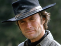 Clint Eastwood w kapeluszu, uśmiechający się z lekko zmarszczonym czołem, w tle zielona natura.