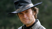 Clint Eastwood w kapeluszu, uśmiechający się z lekko zmarszczonym czołem, w tle zielona natura.