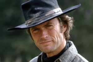 Clint Eastwood w kapeluszu, uśmiechający się z lekko zmarszczonym czołem, w tle zielona natura.