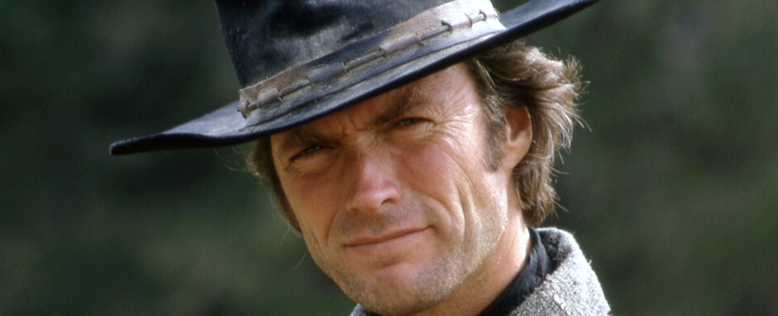 Clint Eastwood w kapeluszu, uśmiechający się z lekko zmarszczonym czołem, w tle zielona natura.