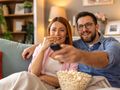 Para siedzi na kanapie, trzymając pilota i jedząc popcorn, oglądając telewizję.
