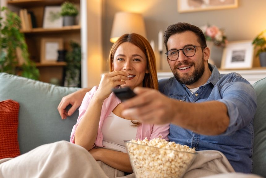 Para siedzi na kanapie, trzymając pilota i jedząc popcorn, oglądając telewizję.