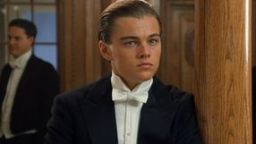 Młody mężczyzna w eleganckim garniturze z białą muszką, stojący obok drewnianej kolumny.