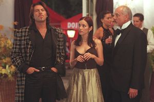 Trzy osoby w eleganckich strojach, w tle dekoracje i inne postacie na przyjęciu.