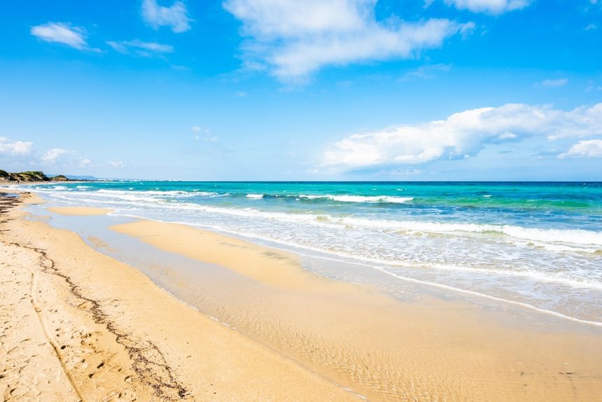 Piaszczysta plaża z falami morskimi, błękitne niebo z białymi chmurami.