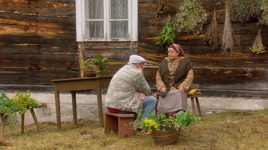 Dwoje ludzi siedzi w ogrodzie przy drewnianym domu, otoczeni roślinami. Rozmawiają ze sobą.