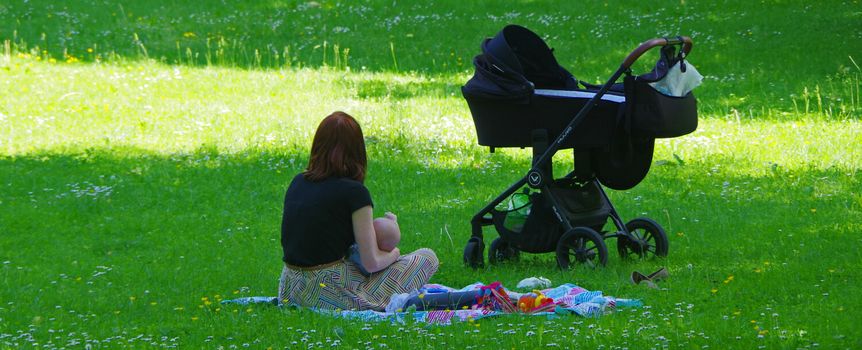 Kobieta w czarnym topie siedzi na kocu z dzieckiem w parku, obok wózek dziecięcy.