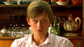 Mężczyzna z blond włosami siedzi w pomieszczeniu z zastawą stołową w tle.