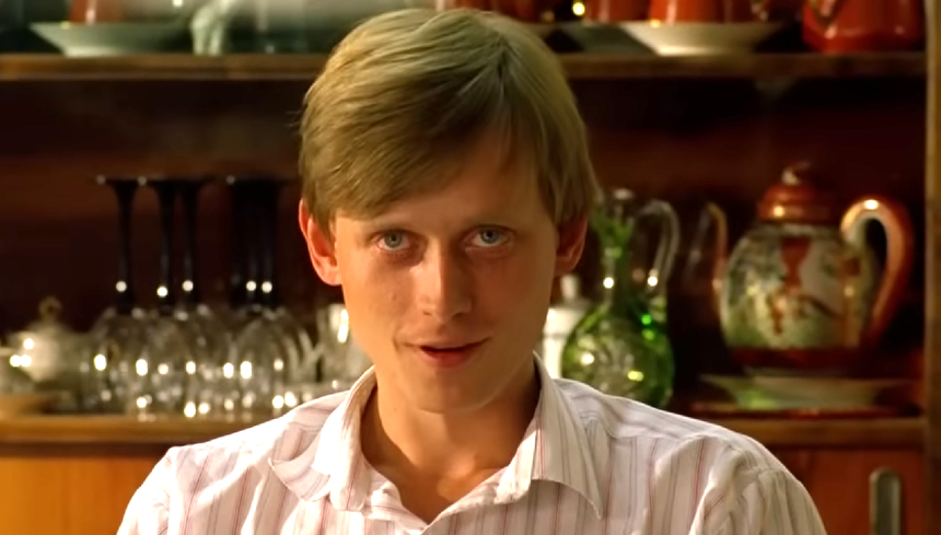 Mężczyzna z blond włosami siedzi w pomieszczeniu z zastawą stołową w tle.
