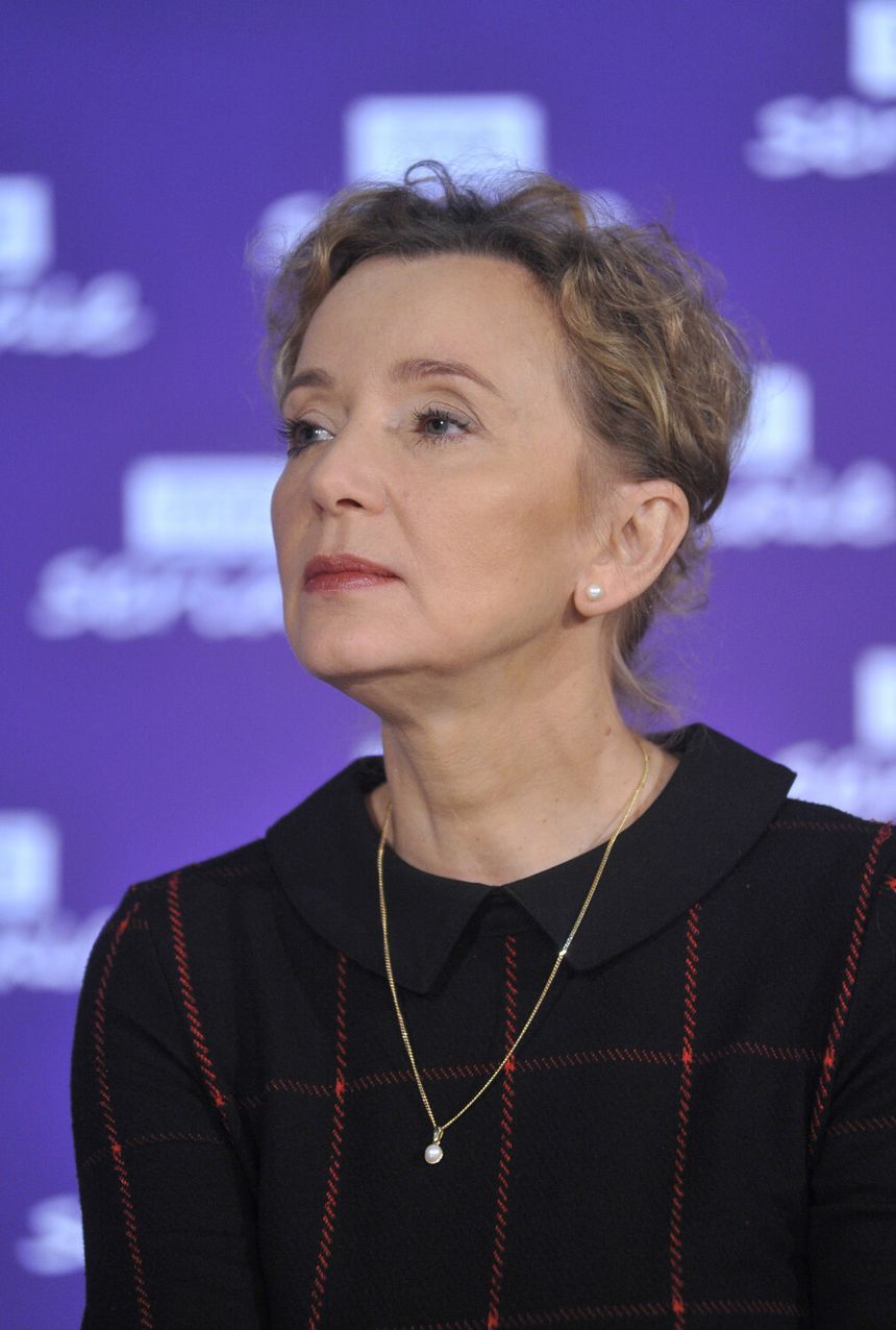 Kobieta z krótkimi włosami, w czarnej sukience, patrzy w górę na tle fioletowego tła.