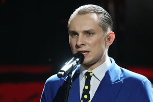 Młody mężczyzna w niebieskim garniturze śpiewa do mikrofonu na scenie.