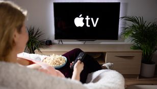 Osoba siedzi na kanapie, trzymając pilota, ogląda ekran z logo Apple TV i je popcorn.