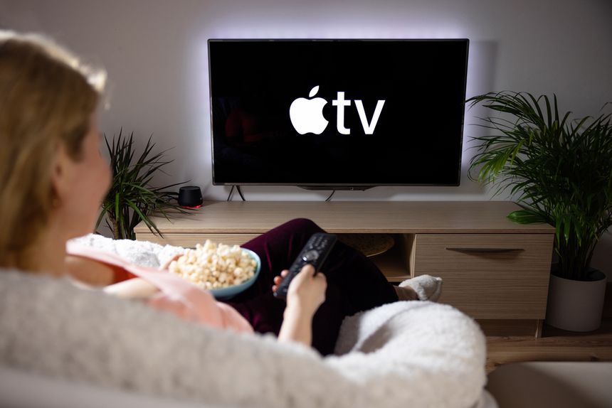 Osoba siedzi na kanapie, trzymając pilota, ogląda ekran z logo Apple TV i je popcorn.