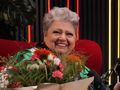 Uśmiechnięta kobieta z szarymi włosami trzyma bukiet kwiat&oacute;w, siedząc na czerwonej kanapie.