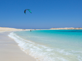 Widok na plażę z turkusową wodą, kitesurfingiem i piaszczystymi wydmami w tle.