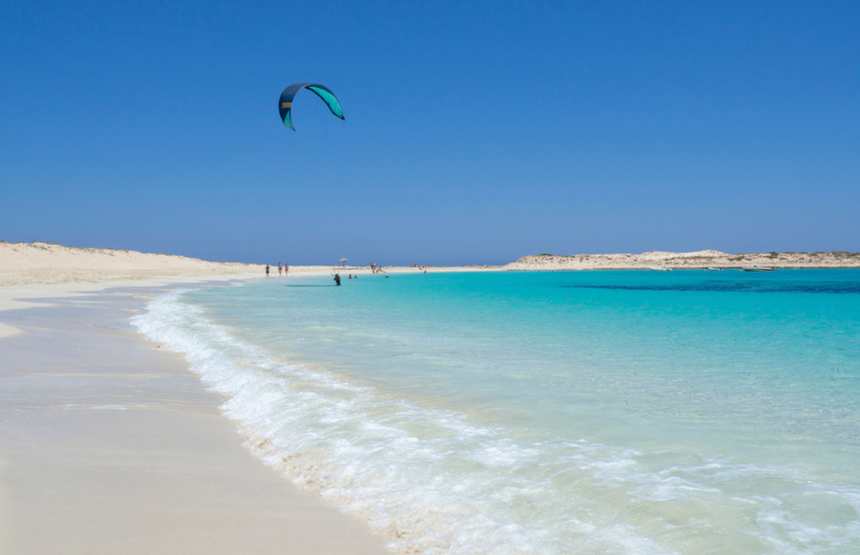Widok na plażę z turkusową wodą, kitesurfingiem i piaszczystymi wydmami w tle.