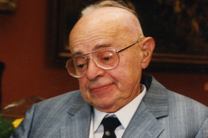 Mężczyzna w okularach, w szarym garniturze, zamyślony, na tle ciemnej ściany.