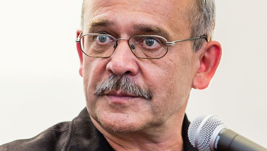 Mężczyzna z okularami, z siwymi włosami i wąsem, patrzy poważnie w stronę obiektywu.