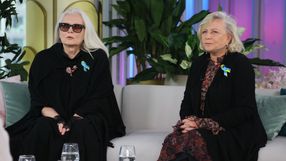 Dwie kobiety w eleganckich czarnych ubraniach siedzą na kanapie, w tle rośliny.