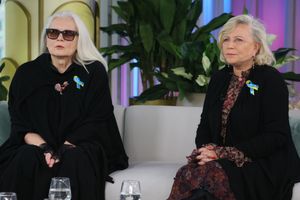 Dwie kobiety w eleganckich czarnych ubraniach siedzą na kanapie, w tle rośliny.