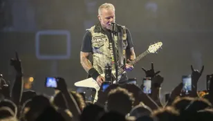 Mężczyzna gra na gitarze i śpiewa do mikrofonu na koncercie. W tle widać tłum fanów.