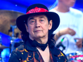Jan Borysewicz w kapeluszu z czerwoną chustą, uśmiechnięty, w otoczeniu instrumentów muzycznych.