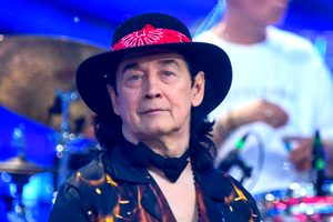 Jan Borysewicz w kapeluszu z czerwoną chustą, uśmiechnięty, w otoczeniu instrumentów muzycznych.