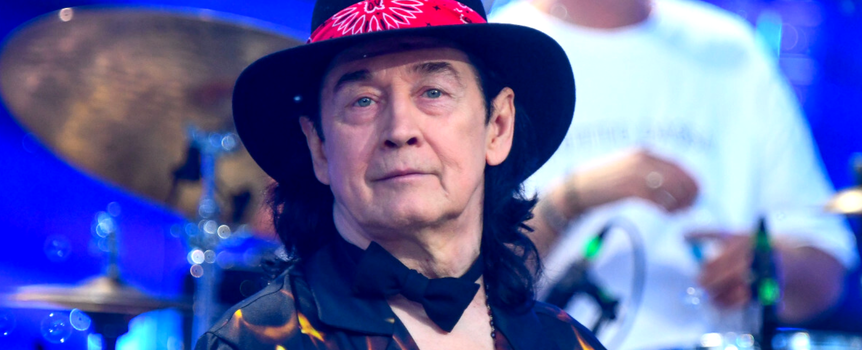Jan Borysewicz w kapeluszu z czerwoną chustą, uśmiechnięty, w otoczeniu instrumentów muzycznych.