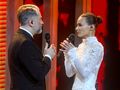Para wykonawców na scenie w eleganckich strojach, trzymających mikrofony. Tło czerwone.