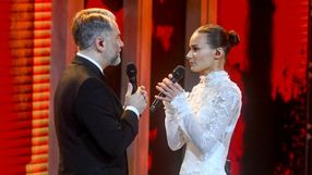 Para wykonawców na scenie w eleganckich strojach, trzymających mikrofony. Tło czerwone.