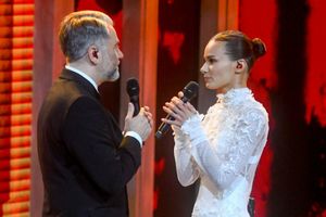 Para wykonawców na scenie w eleganckich strojach, trzymających mikrofony. Tło czerwone.
